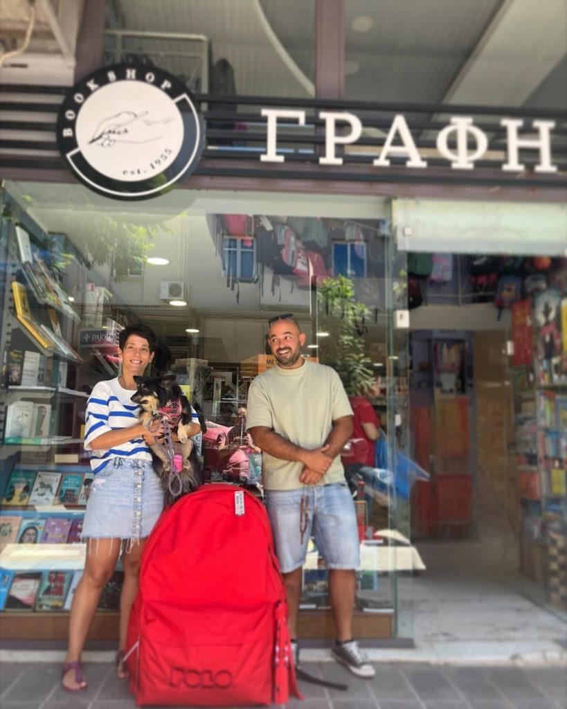 Η τρίτη γενιά της «Γραφής» χαμογελά μπροστά στη βιτρίνα του βιβλιοπωλείου, κάτω από την πινακίδα με το λογότυπο, δίπλα σε μια μεγάλη κόκκινη σχολική τσάντα